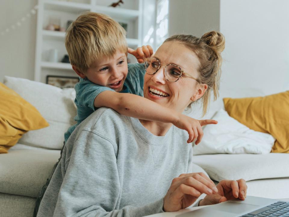 A parent working on a laptop in a bright, cozy living room while a young child leans playfully over their shoulder.